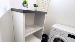 A compact laundry area with a washing machine, a shelf with folded towels, a small potted plant, and a decorative chair.