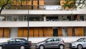 A multi-story building with balconies, parked cars, and a tree in the foreground.