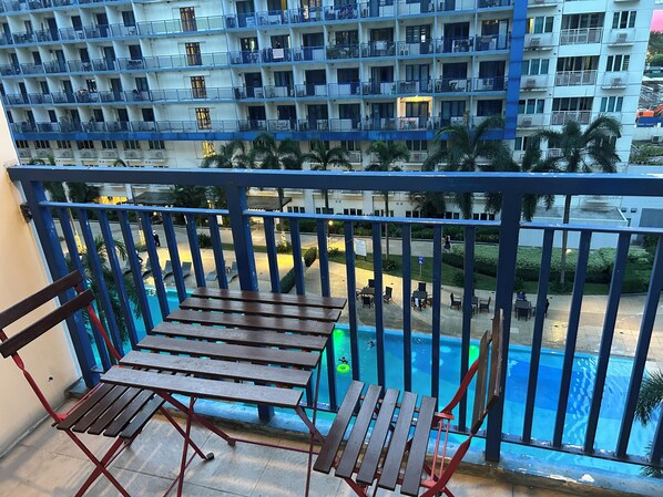 Un balcón con mobiliario de madera, una piscina y vistas a un edificio de varios pisos con balcones.