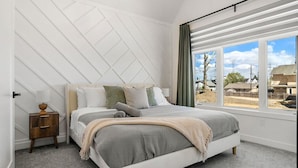 Ein Schlafzimmer mit Bett, Nachttisch, einem Fenster mit Vorhängen und Blick nach draußen.