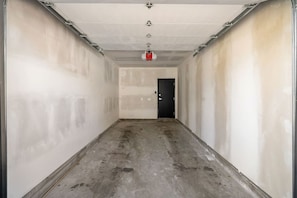 Ein schmaler, unfertiger Flur mit freiliegenden Betonböden und -wänden, einer Tür am Ende und einem an der Decke befestigten Feuerlöscher.