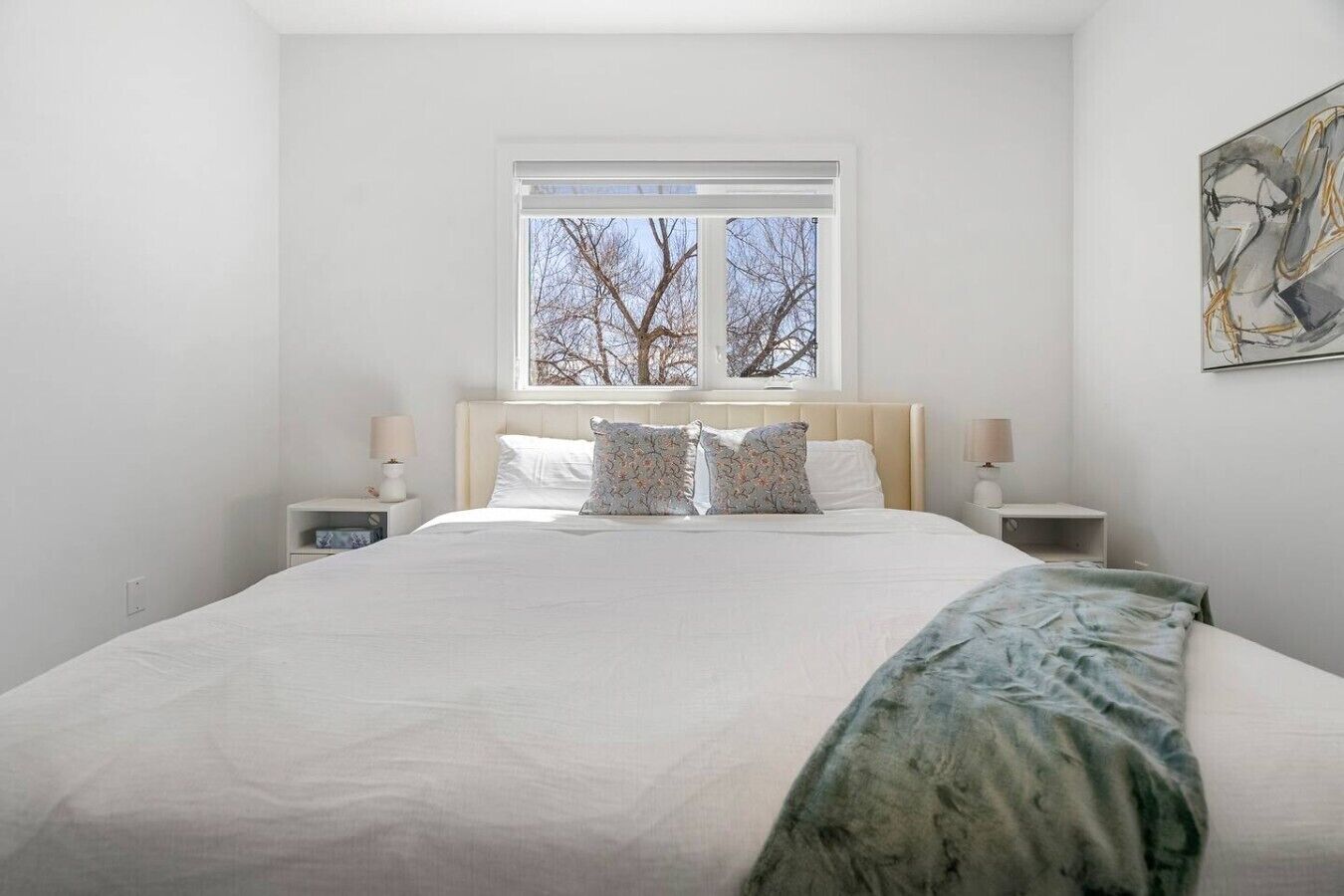 A bedroom with a large bed, two bedside tables, a window with a view, and a piece of abstract art on the wall.