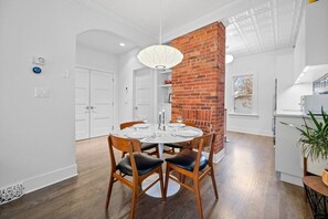 A modern dining area with a brick accent wall, white walls, and a round table set for four. - 2 Bed | Central | Min to Saddledome (Calgary)