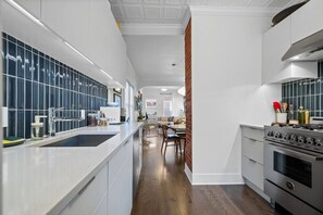 A modern kitchen with white cabinetry, a central island, and a dining area with a brick accent wall. - 2 Bed | Central | Min to Saddledome (Calgary)