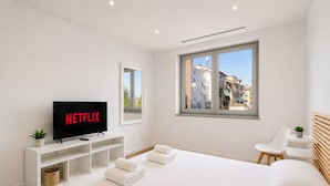 Ein modernes Schlafzimmer mit einem großen Bett, einem Fernseher, der 'NETFLIX' zeigt, einem Fenster mit Blick auf die Stadt und einem kleinen Tisch mit einer Pflanze.