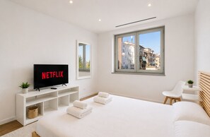 A modern bedroom with a large bed, a TV displaying 'NETFLIX,' a window with a city view, and a small table with a plant. - Design apartment 10 min from downtown with balcony (Torino)