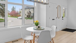 Quarto com mesa redonda, cadeiras brancas, luminária pendente e vista para a rua e casas.