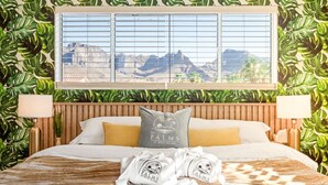 Quarto de hotel com cama, duas almofadas, janela com vista e papel de parede com folhagem tropical.