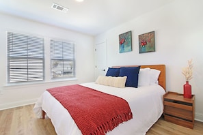 Una camera da letto con un letto, pareti bianche, una coperta rossa, due quadri e un comodino in legno con un vaso.