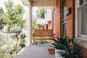 Eine Veranda mit einer hölzernen Bank, einem Blumentopf und einem Backsteinhaus im Hintergrund.