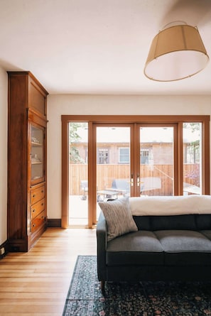 Ein Zimmer mit einer dunklen Couch, einem Holzschrank und einem großen Fenster mit Blick auf einen Hinterhof.