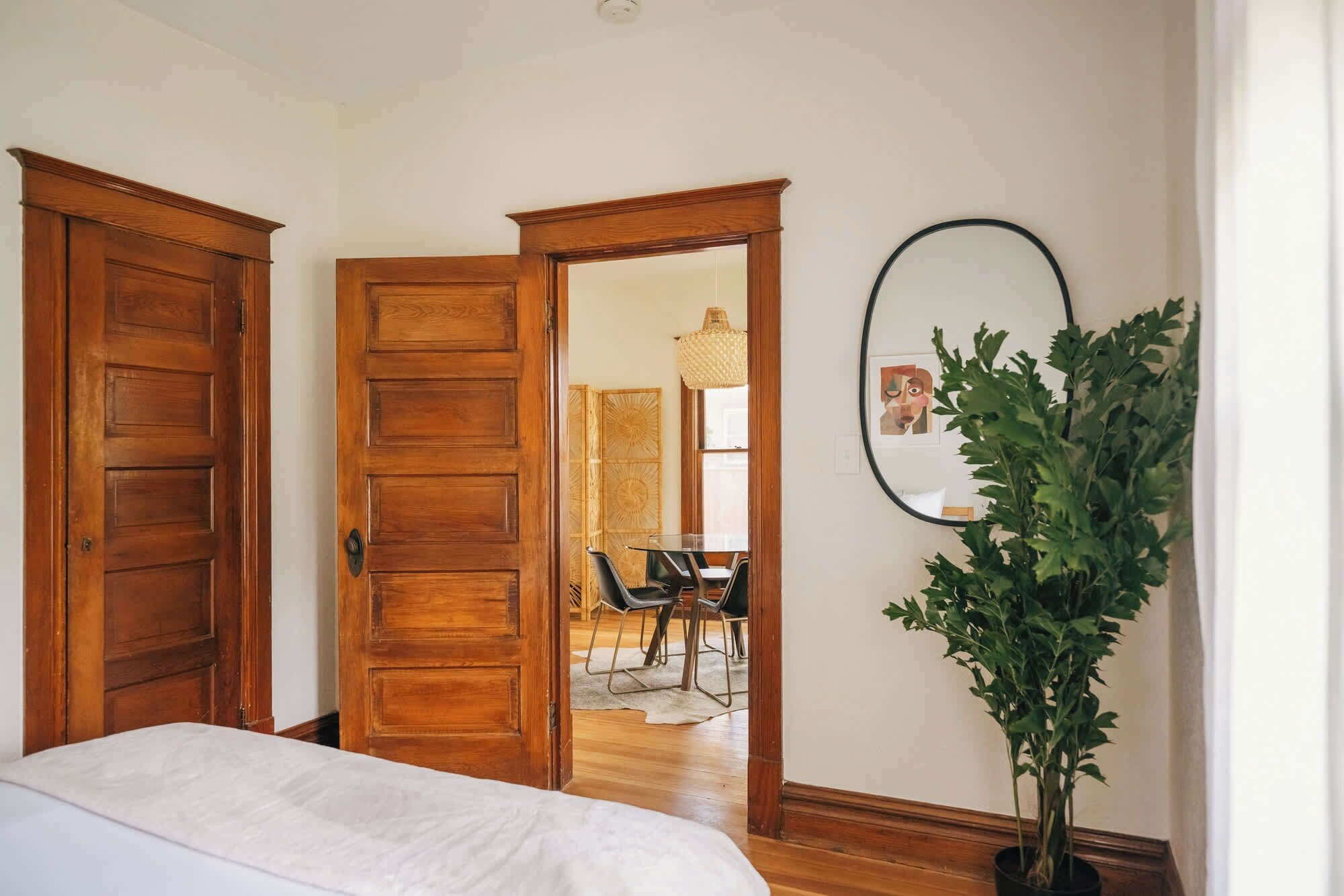 A bedroom with a bed, wooden doors, a mirror, a potted plant, and a dining area in the background.