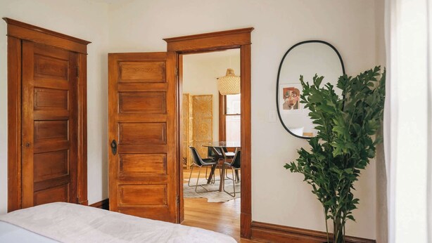 Une chambre à coucher avec un lit, des portes en bois, un miroir, une plante en pot et, au fond, un coin repas.