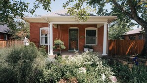 Une maison à deux étages, construite en briques, avec un porche couvert, une cour avant agrémentée de diverses plantes et une clôture en bois.