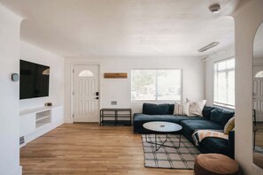 Ein Wohnzimmer mit einem dunkelgrünen Sofa, einem runden Couchtisch, einem gemusterten Teppich, einem an der Wand montierten Fernseher und einer Tür mit Fenster.