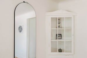 Ein Zimmer mit einem weißen Schrank mit Glastüren, einem spiegeln Spiegel mit schwarzem Rahmen und einem kleinen Bild in einem Rahmen auf dem Schrank.