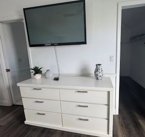 A white dresser with drawers, a flat-screen TV mounted above it, a small potted plant, and a decorative vase on the dresser. - Spacious 4-bedroom house in delightful Cape Coral with AC (Cape Coral)