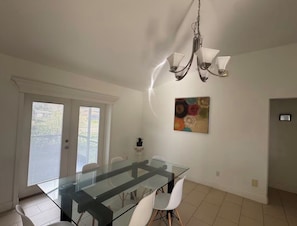 A dining area with a glass table, white chairs, a chandelier, and a painting on the wall. - Spacious 4-bedroom house in delightful Cape Coral with AC (Cape Coral)