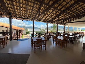 Un área de comedor al aire libre con mesas y sillas de madera, techo cubierto y vista al paisaje.