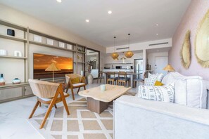 Ein modernes Wohnzimmer mit einer Couch, Sesseln, einem Couchtisch und einem Essbereich. Im Hintergrund ist eine Küche zu sehen.