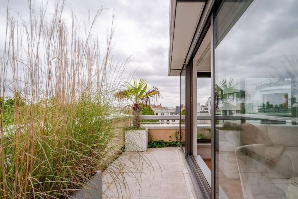 Un balcon moderne agrémenté de hautes herbes, d’un palmier et d’une porte vitrée donnant sur un espace de vie avec un canapé.