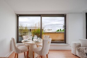 A dining area with a round table and chairs, a view through large windows, and a balcony with plants. - Elysee's Residence / Luxury 1 Bed Serviced Apartment (Paris)