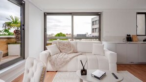 Ein modernes Wohnzimmer mit einem weißen Sofa, einem kleinen runden Tisch mit einer Fernbedienung und Blick auf die Stadt durch große Fenster.