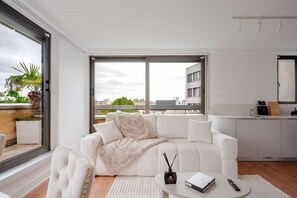 A modern living room with a white sofa, a small round table with a remote control, and a view of the city through large windows. - Elysee's Residence / Luxury 1 Bed Serviced Apartment (Paris)