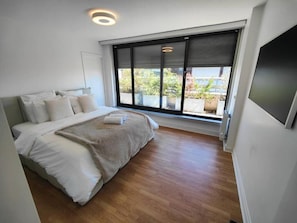 A modern bedroom with a large bed, a view of greenery outside, and a flat-screen TV on the wall. - Elysee's Residence / Luxury 1 Bed Serviced Apartment (Paris)