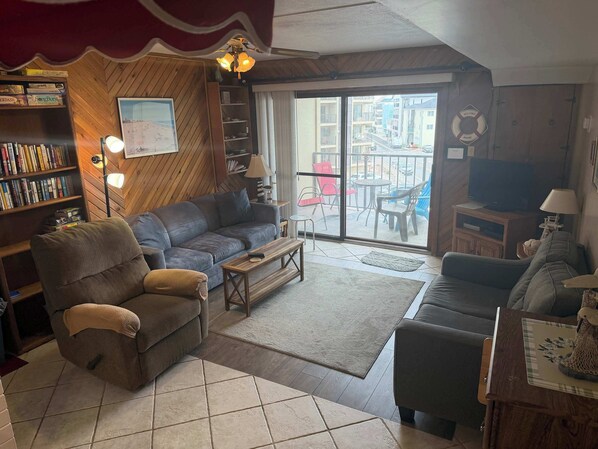 Un salon comprenant un canapé, un fauteuil et une table basse, une bibliothèque, un téléviseur et un balcon aménagé avec du mobilier d’extérieur.