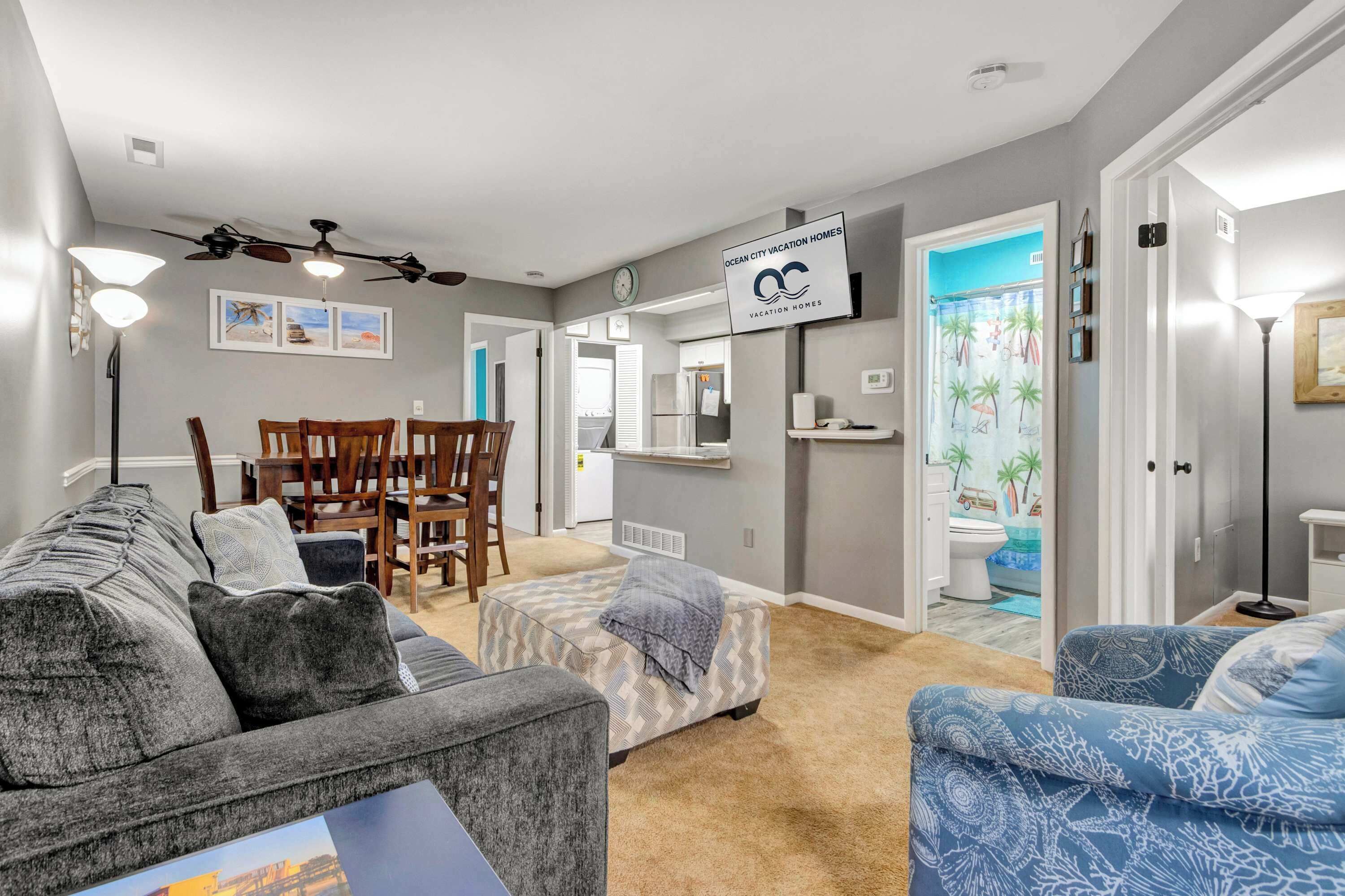 A living room with a grey sofa, ottoman, and blue patterned chair, a dining area with a table and chairs, a kitchen with a refrigerator, and a bathroom with a shower curtain.