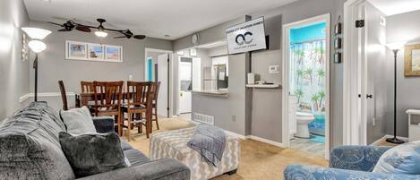 A living room with a grey sofa, ottoman, and blue patterned chair, a dining area with a table and chairs, a kitchen with a refrigerator, and a bathroom with a shower curtain.