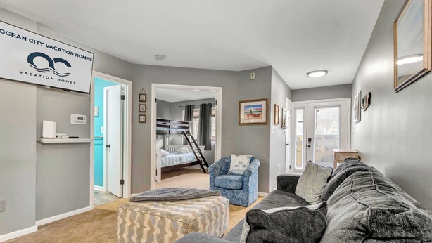 A living room with a gray couch, ottoman, and a blue chair. There is a bed with a ladder in the background. - Sherwood 1C (Ocean City)