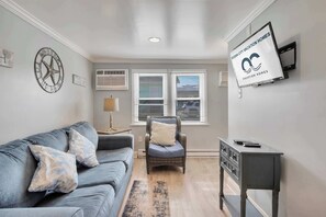 Ein Wohnzimmer mit grauem Sofa, einem Korbstuhl, einem kleinen Tisch und einem angebrachten Fernseher, der 'Ocean City Vacation Homes' zeigt.