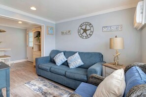 Ein Wohnzimmer mit einem blauen Sofa, einer runden Wanduhr und Wanddekorationen, darunter Schilder mit den Aufschriften 'BEACH' und 'SEASIDE'.