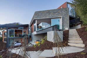 Uma casa moderna com fachada de pedra, um quarto visível com cama, e uma escadaria que leva a um segundo andar.