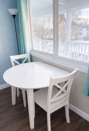 A white round table with two chairs, a window with a view of a white fence and trees, and a floor lamp.