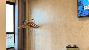 Ein modernes Zimmer mit Holzboden, einem an der Wand montierten Fernseher, einem Metall-Garderobeständer, einem Klappstuhl und einem Holzregal mit einer Kakteenpflanze.