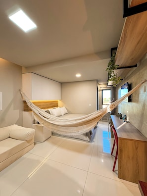 A modern bedroom with a hammock bed, a sofa, a wooden desk, and a view of the outdoors. - Studio Aconchegante Território Serra da Capivara (São Raimundo Nonato)
