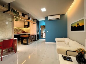 A modern living room with a sofa, a dining area with chairs, a kitchen with a microwave, and an air conditioning unit on the wall.