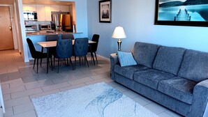 Een moderne woonkamer met een grijze bank, een eethoek met een tafel en stoelen, een keuken op de achtergrond en een ingelijst schilderij aan de muur.