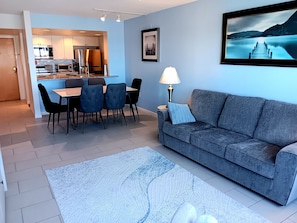 Una sala de estar moderna con un sofá gris, una zona de comedor con una mesa y sillas, una cocina al fondo y un cuadro en la pared.