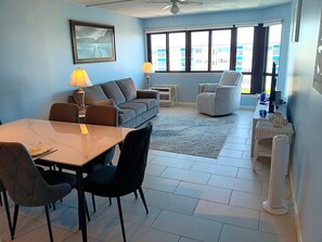Een woonkamer met een bank, een eettafel met stoelen, een staande ventilator en een raam met uitzicht op gebouwen.