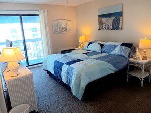 A bedroom with a bed, two bedside tables with lamps, a window with curtains, a painting on the wall, and a view of buildings outside. - Beautiful Brigantine Beach Beach Block Condo--2 Bed--2 Bath (Brigantine)
