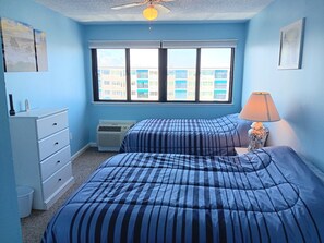 Une chambre avec deux lits, une commode, un radiateur, une fenêtre donnant sur des immeubles et un ventilateur de plafond.