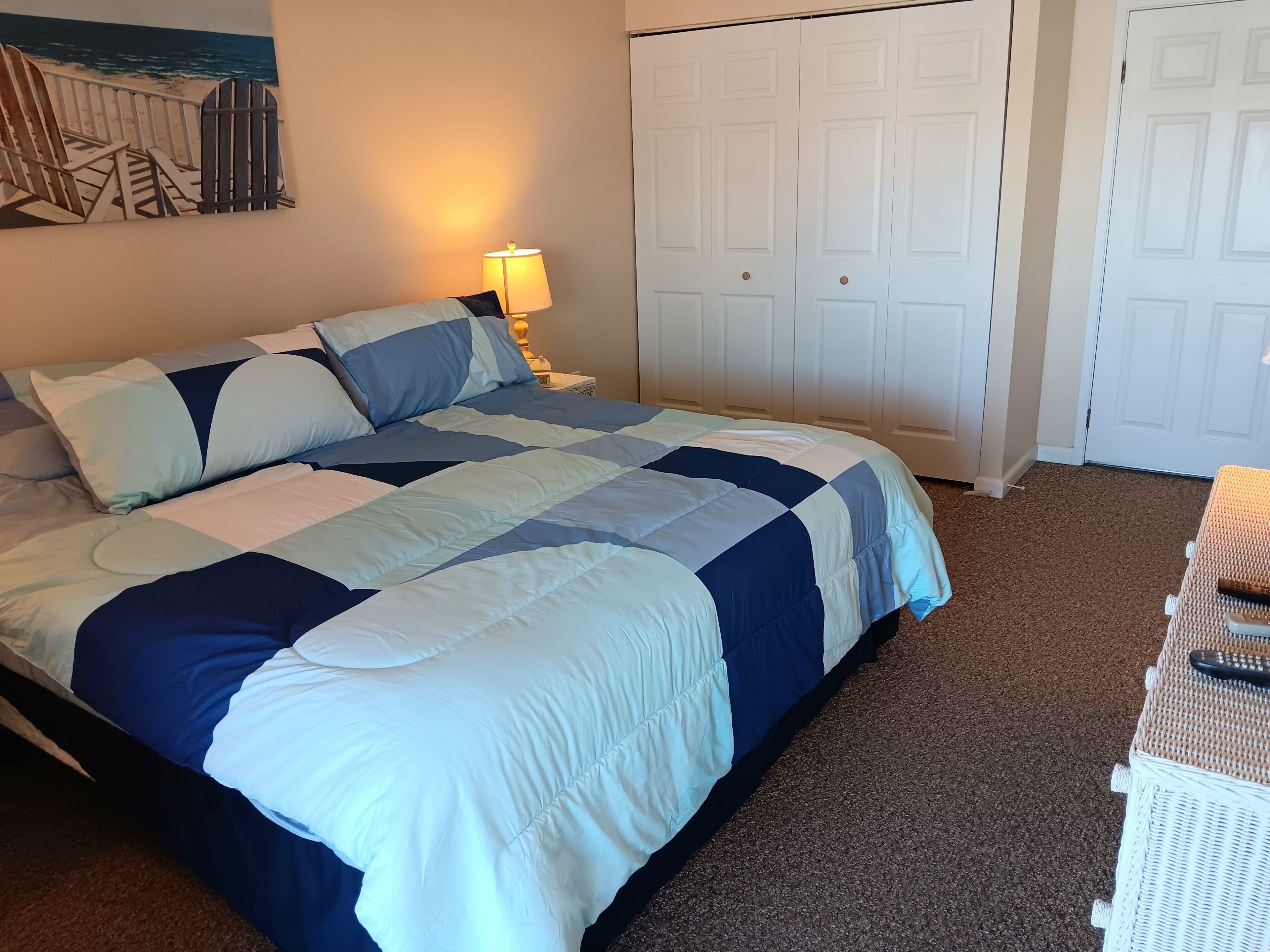 A bed with a blue and white quilt, a nightstand with a lamp, a closet with white doors, and a beach-themed artwork on the wall.