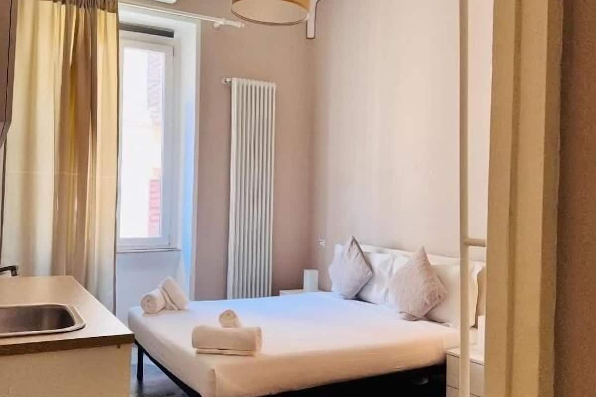 Une chambre d’hôtel avec un lit, un petit bureau, un radiateur et une fenêtre avec des rideaux.