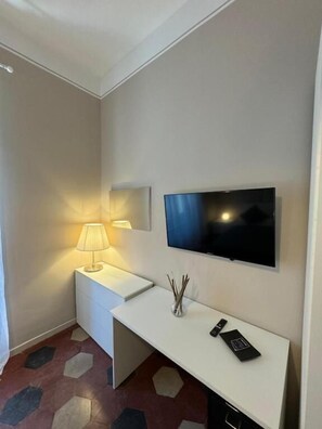 Ett hotellrum med en platt-TV som är monterad på väggen, ett vitt sängbord med en lampa och en dekorativ vas.
