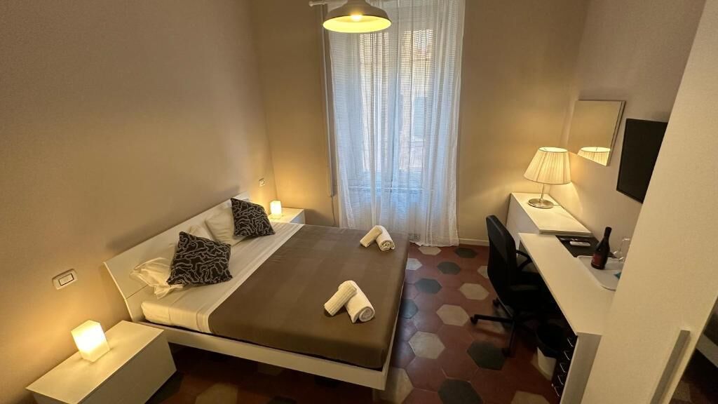 A hotel room with a bed, bedside table, lamp, desk, chair, and a window with sheer curtains.