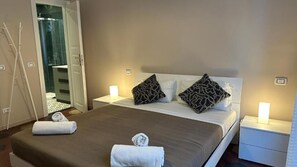 A hotel room with a bed, two bedside tables, a nightstand, a bathroom visible through an open door, and a towel on the bed. - VATICANO ROMA GUESTHOUSE (Rome)